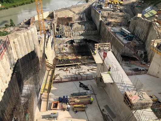 Large construction site next to a river, where several construction workers and construction equipment are working in a deep excavation pit.