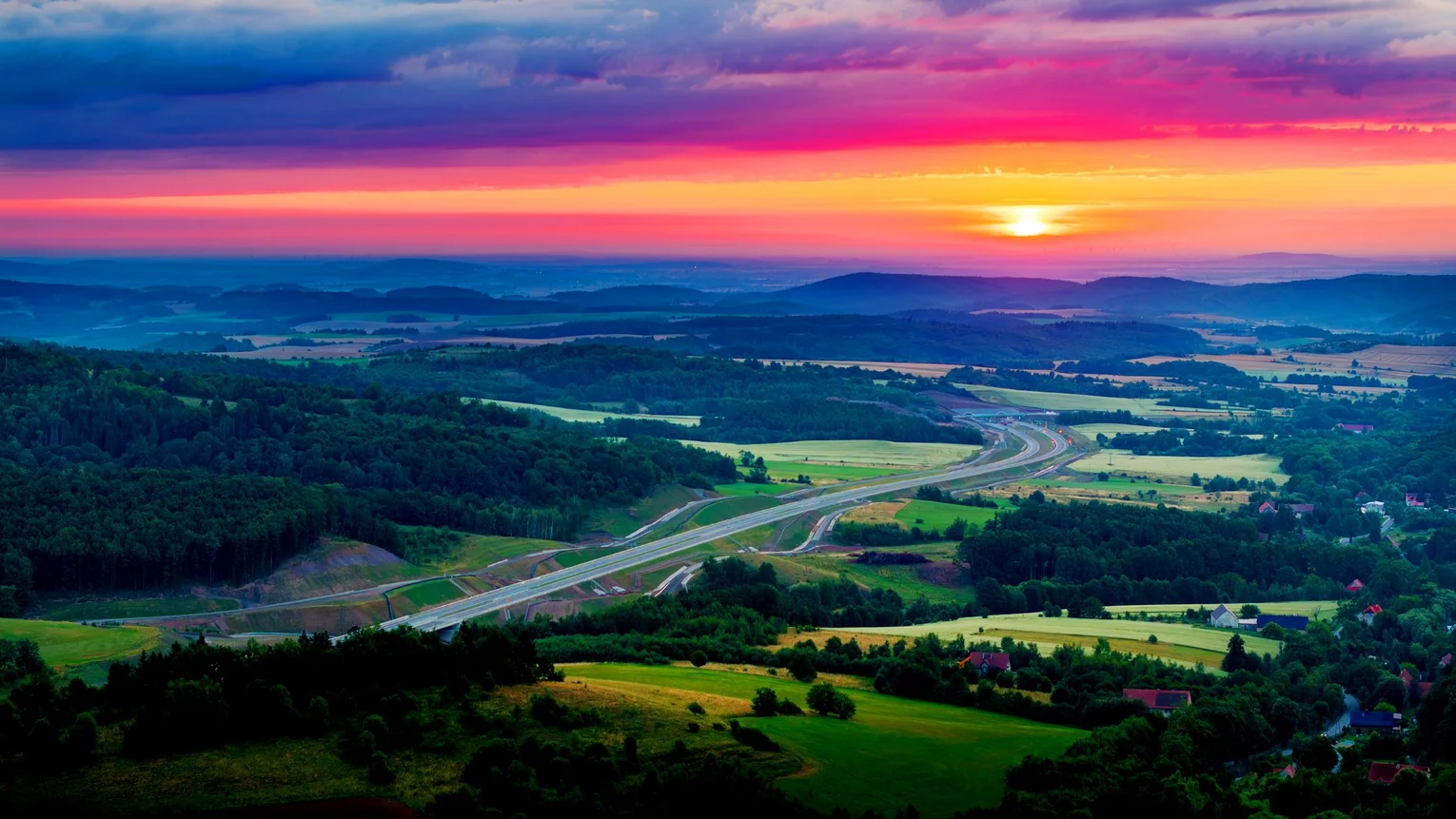 Landscape with meadows and forests through which a road leads, sunset in the background