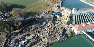 An aerial view of a large construction site next to a river, where several construction machines and cranes are working.